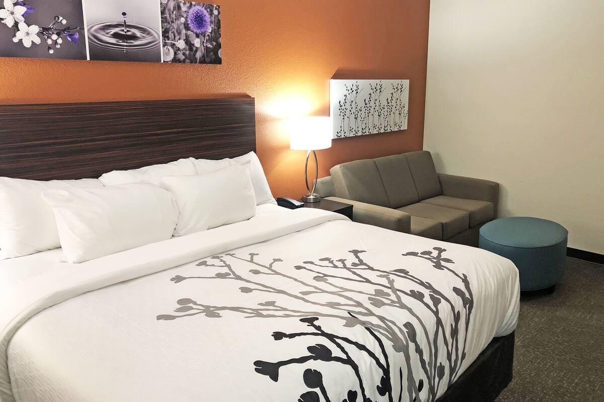 A hotel room with a large bed, a sofa, a round ottoman, a nightstand with a lamp, and wall art.