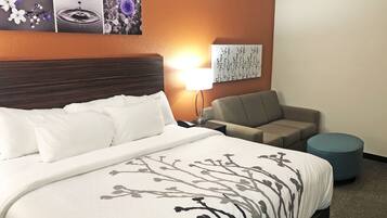 Habitación de hotel con una cama grande, un sofá, un otoman redondo, una mesita de noche con lámpara y decoración en la pared.