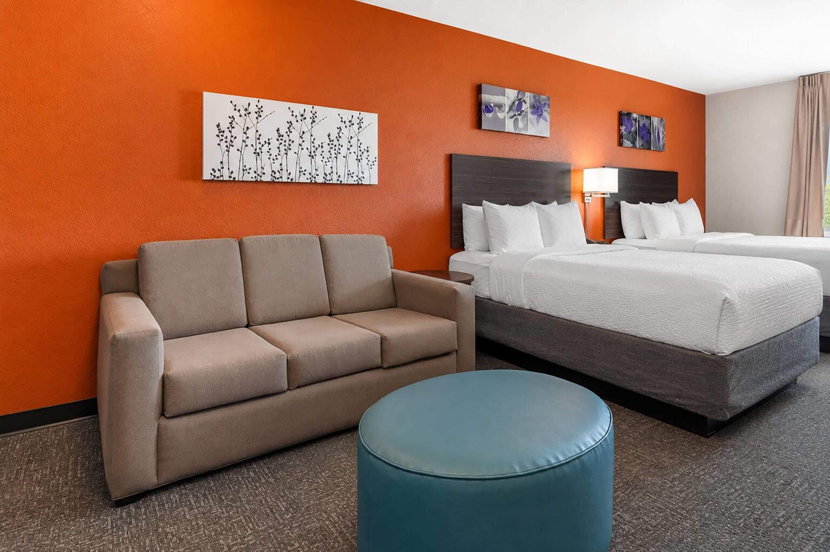 A hotel room with a beige sofa, a blue ottoman, a bed with white bedding, and two wall-mounted paintings.