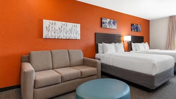 A hotel room with a beige sofa, a blue ottoman, a bed with white bedding, and two wall-mounted paintings.
