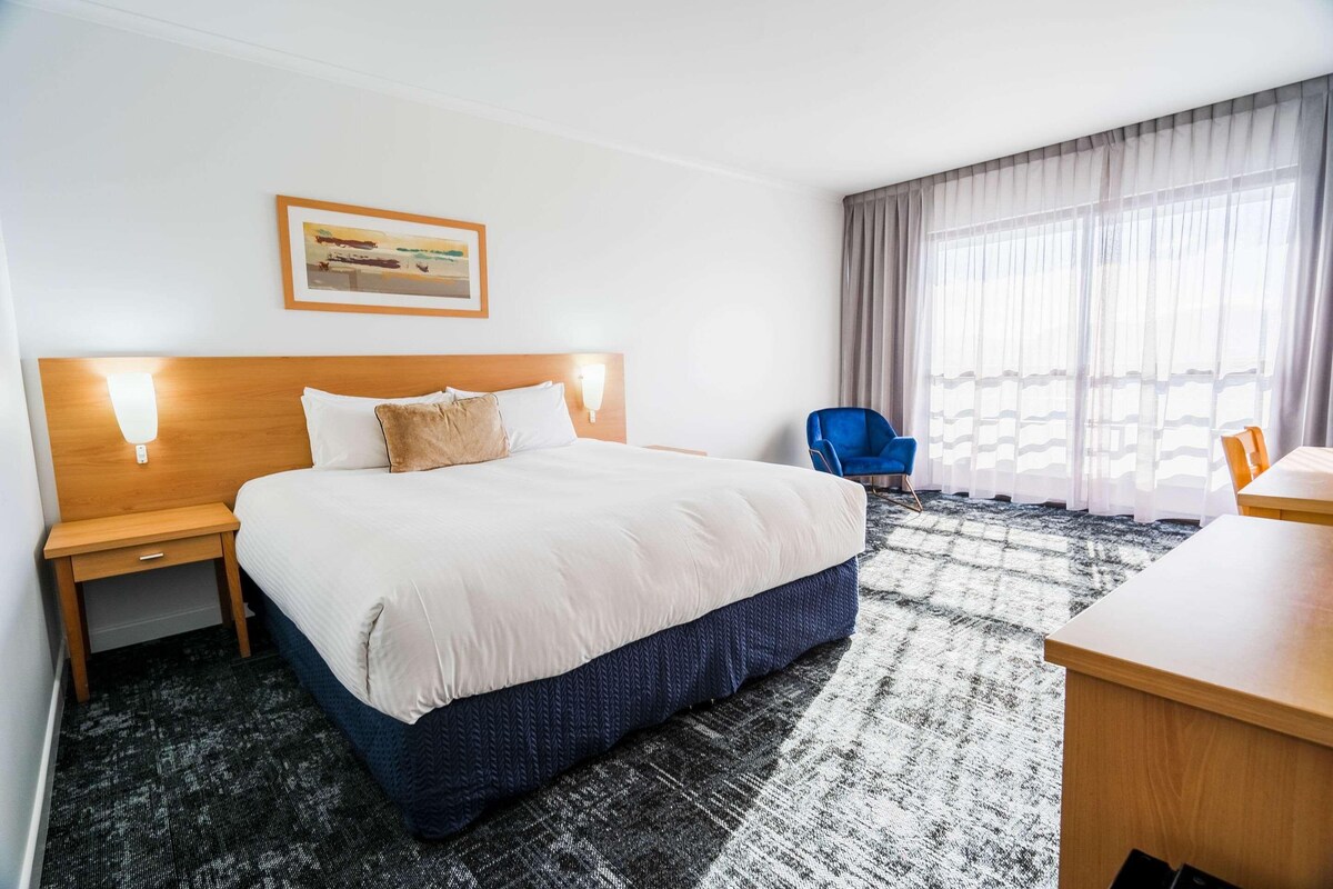 A hotel room with a large bed, two bedside tables, a desk, a chair, and a window with curtains.