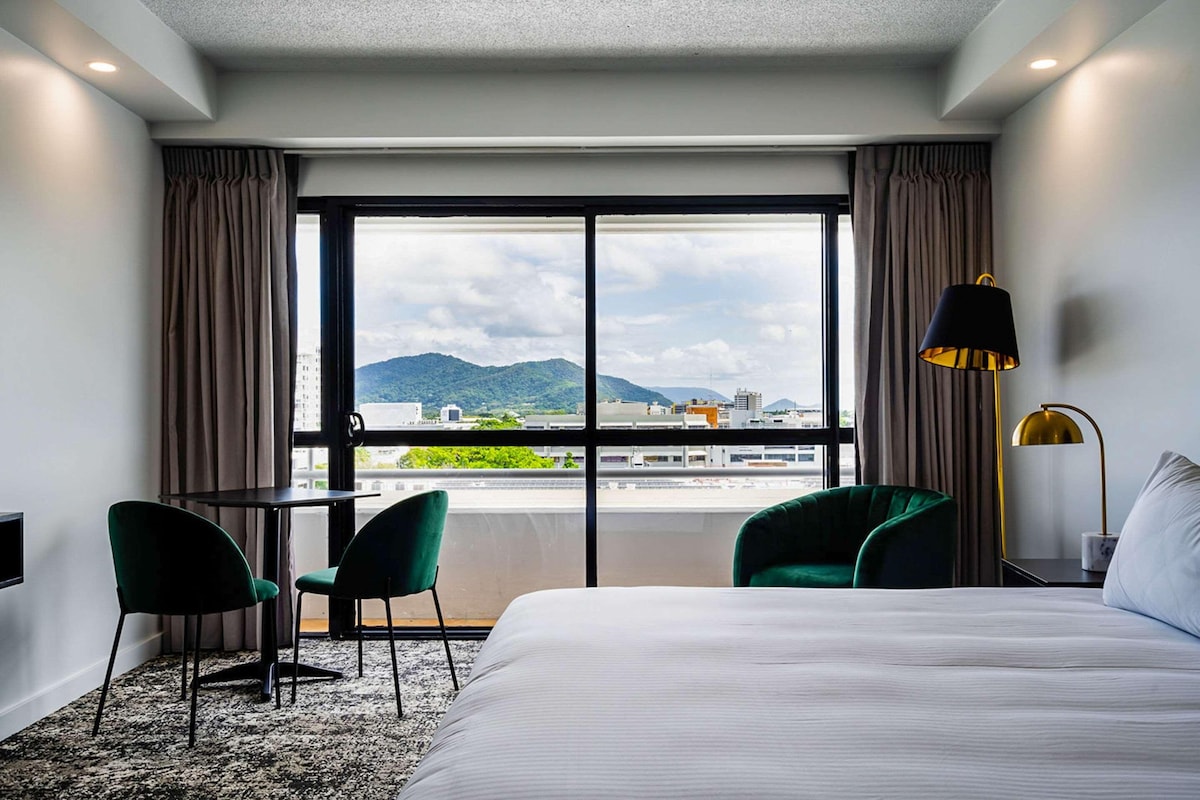 A hotel room with a bed, two green chairs, a small table, a lamp, and a large window with a city and mountains view.