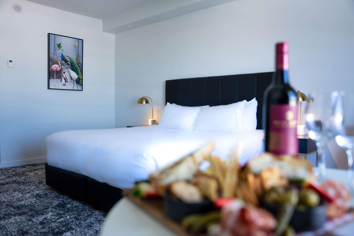 A hotel room with a large bed, a nightstand with a lamp, and a dining table with food and a bottle of wine.
