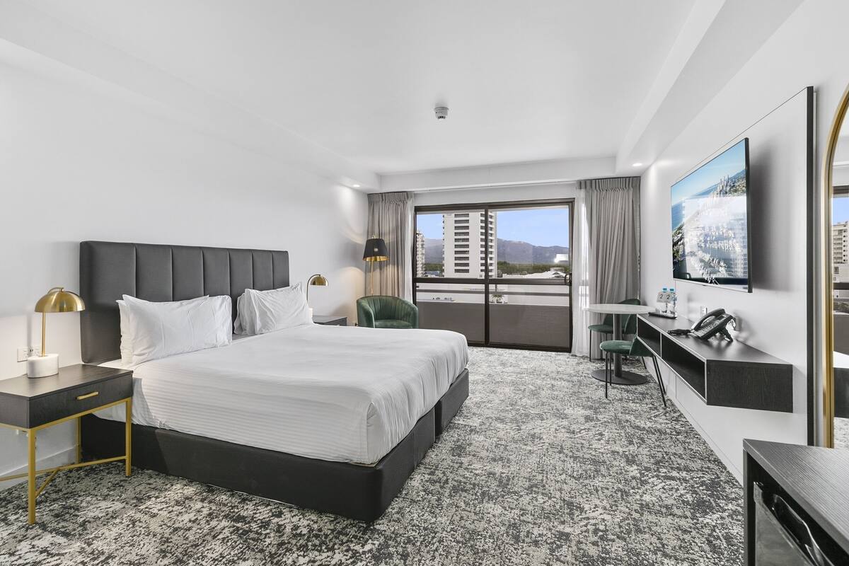 A modern hotel room with a large bed, a desk, a chair, a TV, and a balcony with a city view.