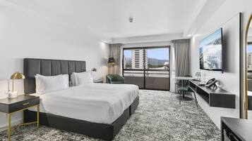 A modern hotel room with a large bed, a desk, a chair, a TV, and a balcony with a city view.