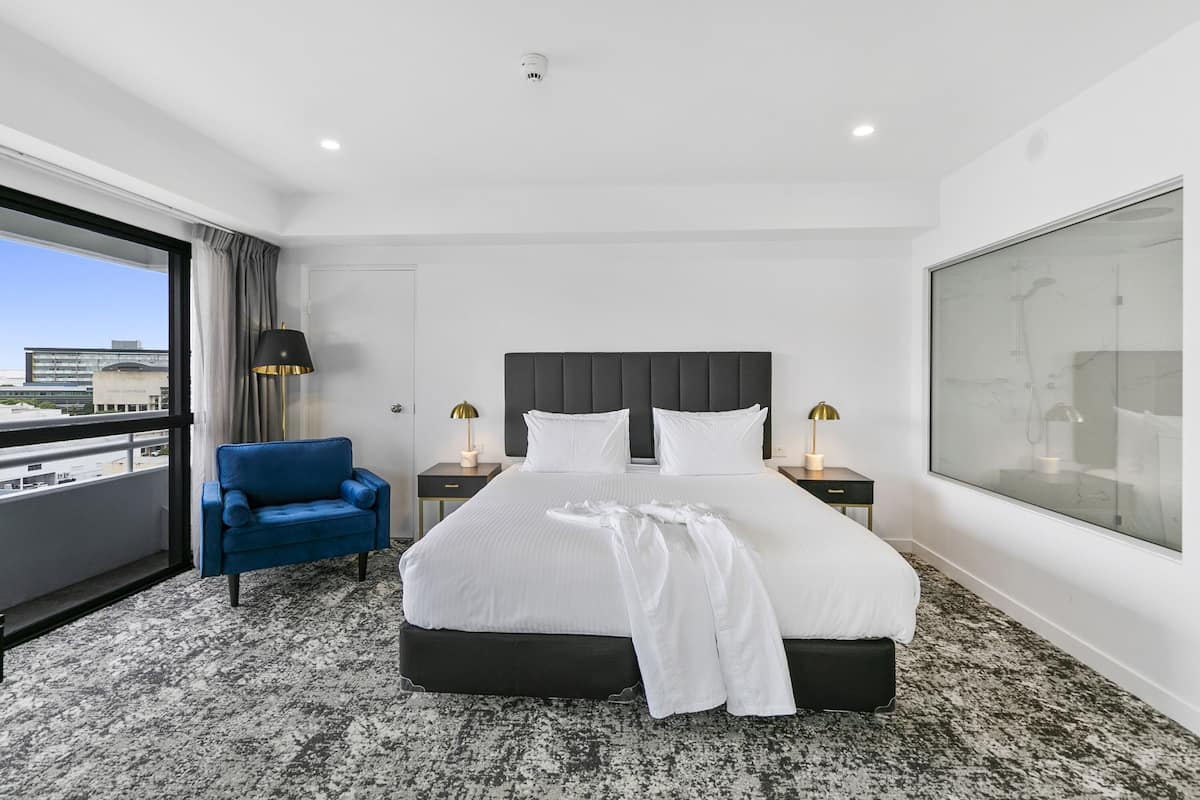 A hotel room with a large bed, a blue armchair, two bedside tables with lamps, a large window with a view of buildings, and a marble-patterned carpet.