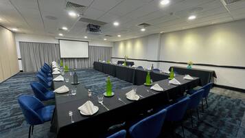 Un salón de conferencias con una larga mesa, sillas azules y una pantalla de proyector.