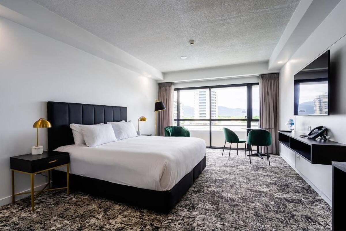 A modern hotel room with a large bed, a desk with a lamp, a chair, and a TV.