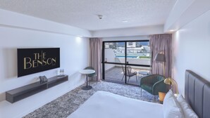 A hotel room with a large bed, a TV mounted on the wall, a small table, and a view of the pool and buildings outside. - The Benson Hotel (Cairns)