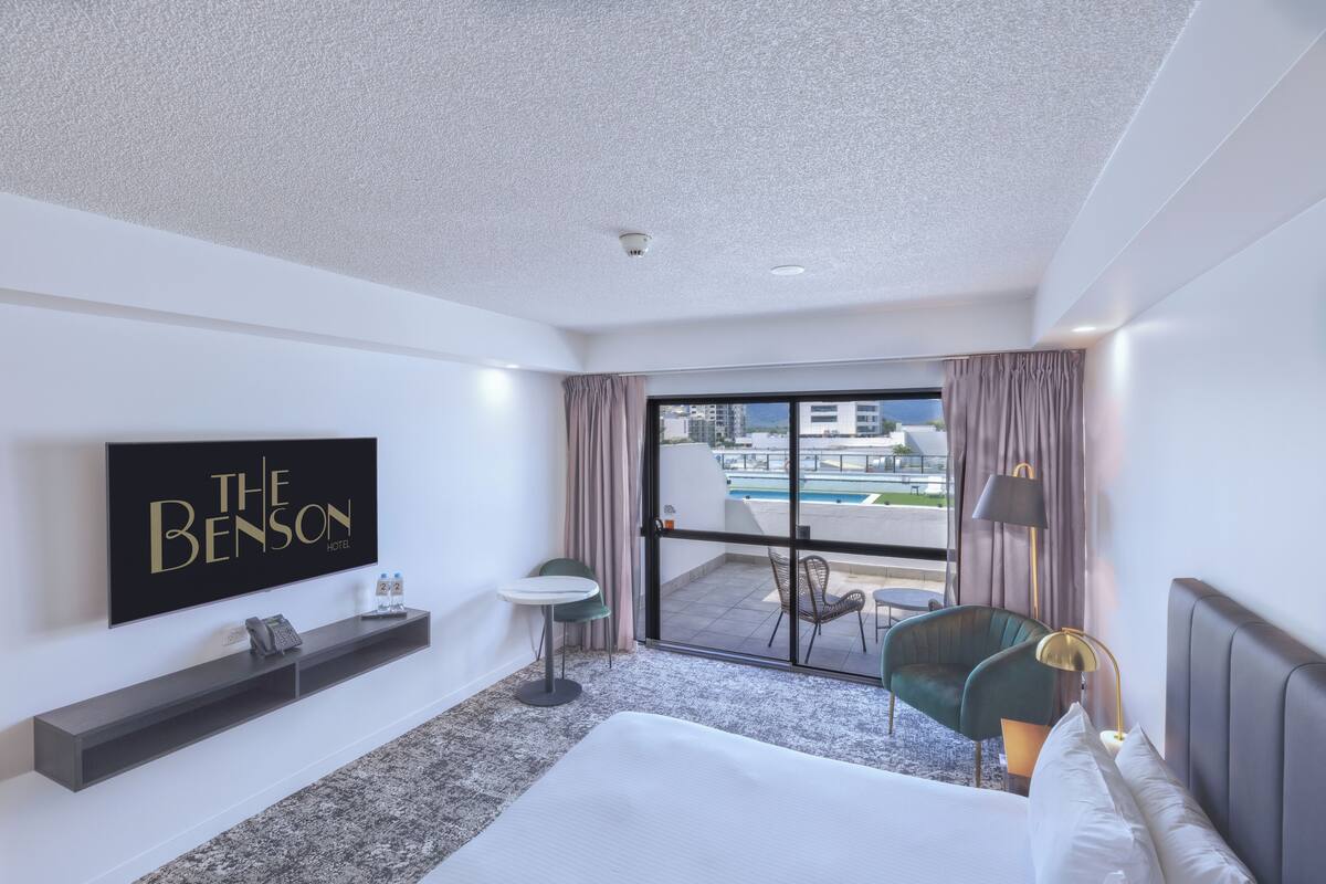 A hotel room with a large bed, a TV mounted on the wall, a small table, and a view of the pool and buildings outside.