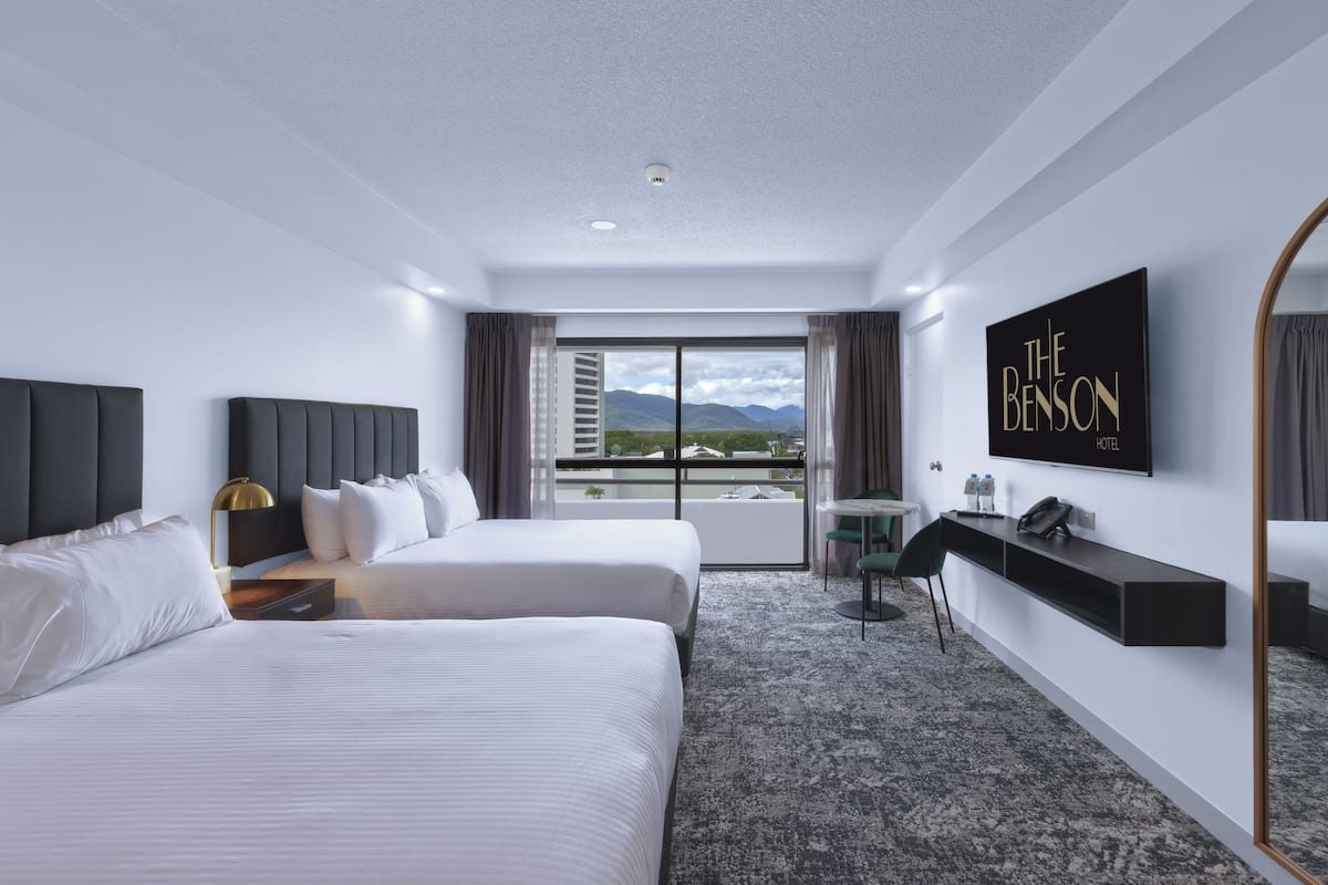 A hotel room with two beds, a desk, a chair, a TV, and a view of mountains.