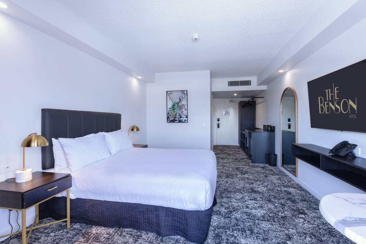 A modern hotel room with a large bed, a desk with a lamp, a mirror, and a wall-mounted TV.