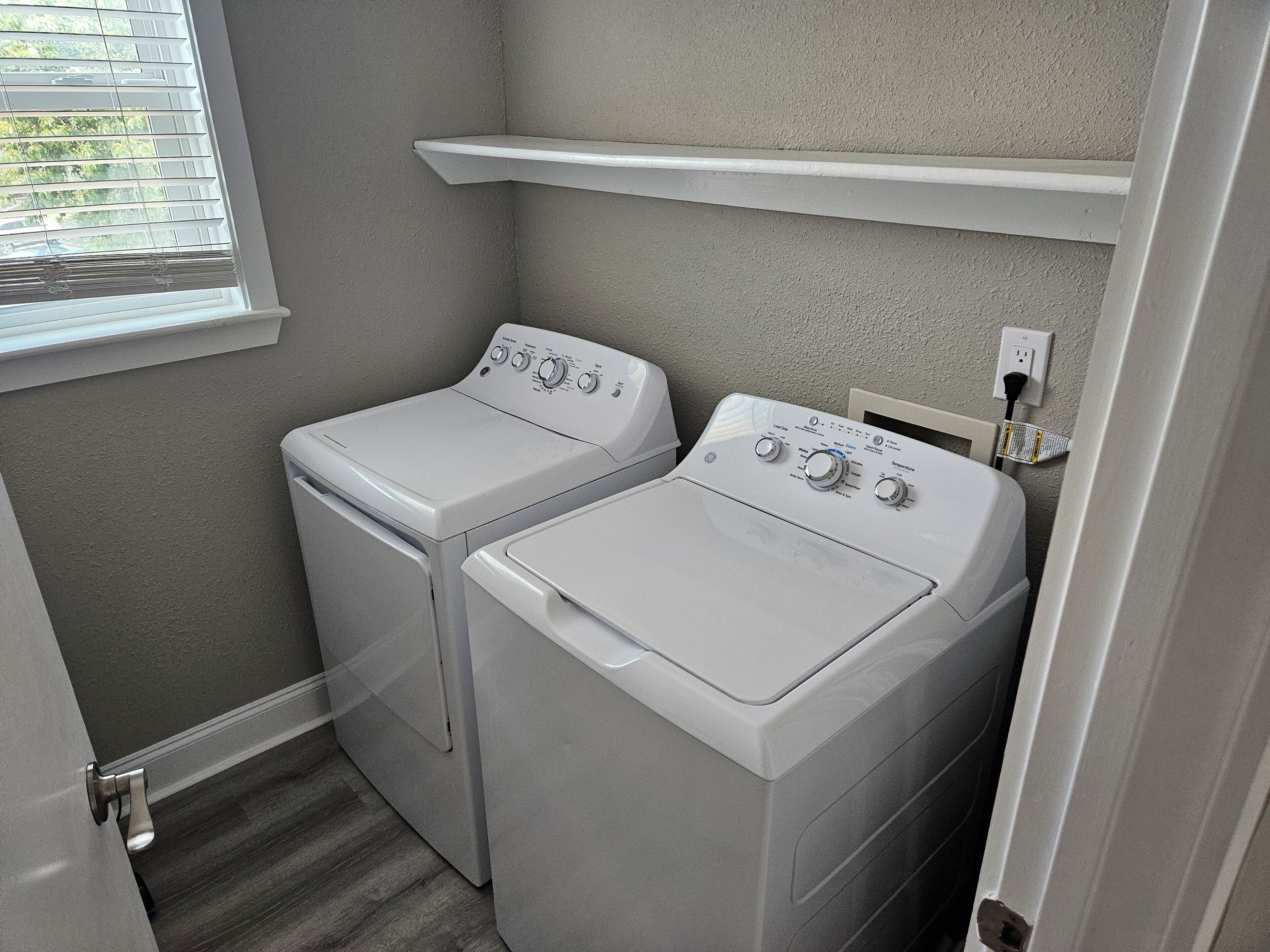 A laundry room with two white front-loading washing machines, a window with blinds, and a shelf above the machines.