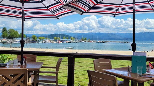 A terrace with tables and chairs under striped umbrellas, overlooking a marina with boats and a beach. - Wooded, peaceful setting 15 minutes from  the Lake! (Sandpoint)