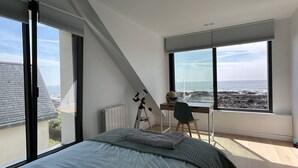 A bedroom with a large bed, a desk, and a chair, overlooking a rocky coastline. - Maison Avec Vue Panoramique sur L'océan (BATZ SUR MER)