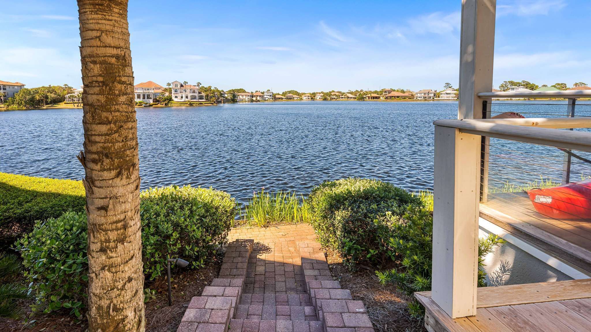 A waterfront property with a paved pathway leading to a deck overlooking a lake, surrounded by lush greenery and residential buildings in the background.