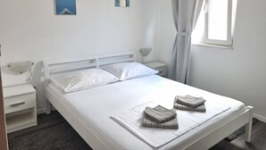 A neatly made bed with white linens, a bedside table with a lamp, and two small wall art pieces. - Apartment Maya 1 (Marušici)