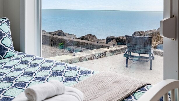 A bedroom with a large window overlooking a beach, a bed with a patterned bedspread, a chair, and a small table with a towel. - Lakefront Retreat + Hot Tub • Sunset Views • Point Pelee (Leamington)