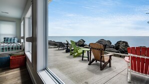 A room with a large window overlooking a rocky shoreline and the sea. There is a bed with patterned bedding and a small table with chairs. - Lakefront Retreat + Hot Tub • Sunset Views • Point Pelee (Leamington)
