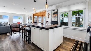 A modern kitchen with a large island, pendant lighting, and large windows offering views of the outdoors. - Lakefront Retreat + Hot Tub • Sunset Views • Point Pelee (Leamington)