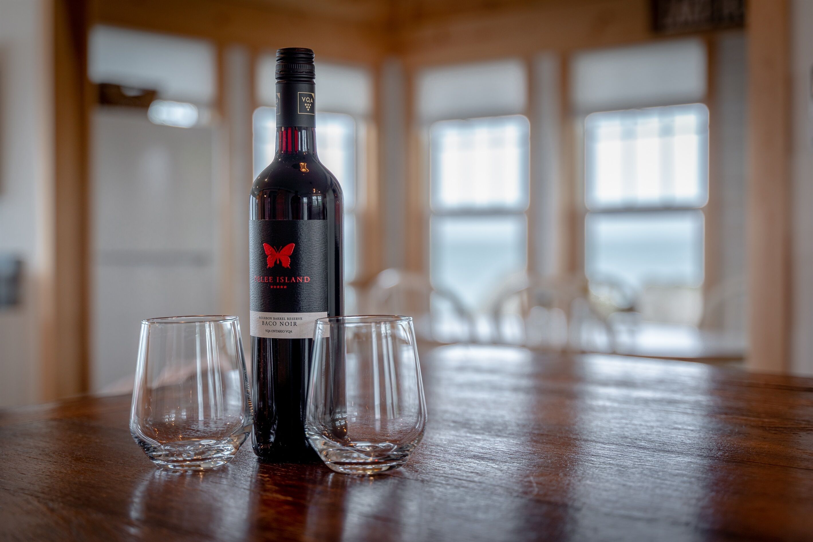 Une bouteille de vin rouge, accompagnée de deux verres, est posée sur une table en bois dans une pièce dotée de grandes fenêtres.