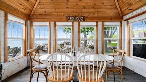 A wooden-paneled room with a round table and four chairs, a sign reading "LAKE HOUSE", and large windows overlooking a rocky shoreline. - Lakefront Retreat + Hot Tub • Sunset Views • Point Pelee (Leamington)