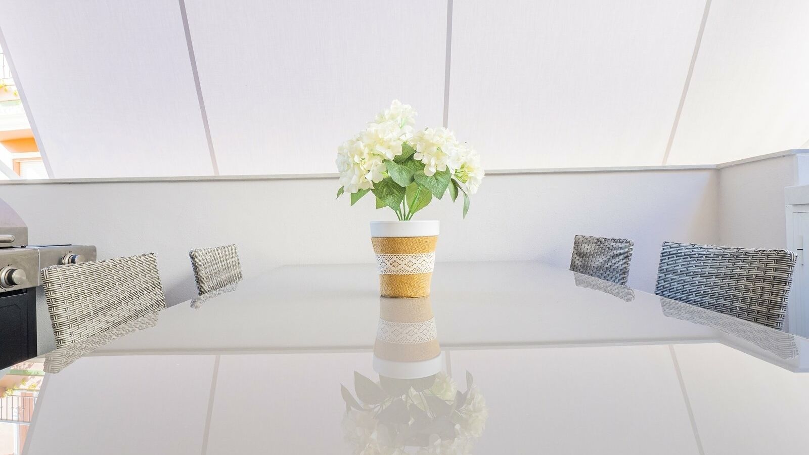 A modern dining area with a glass table, white cushions, and a vase with white flowers.