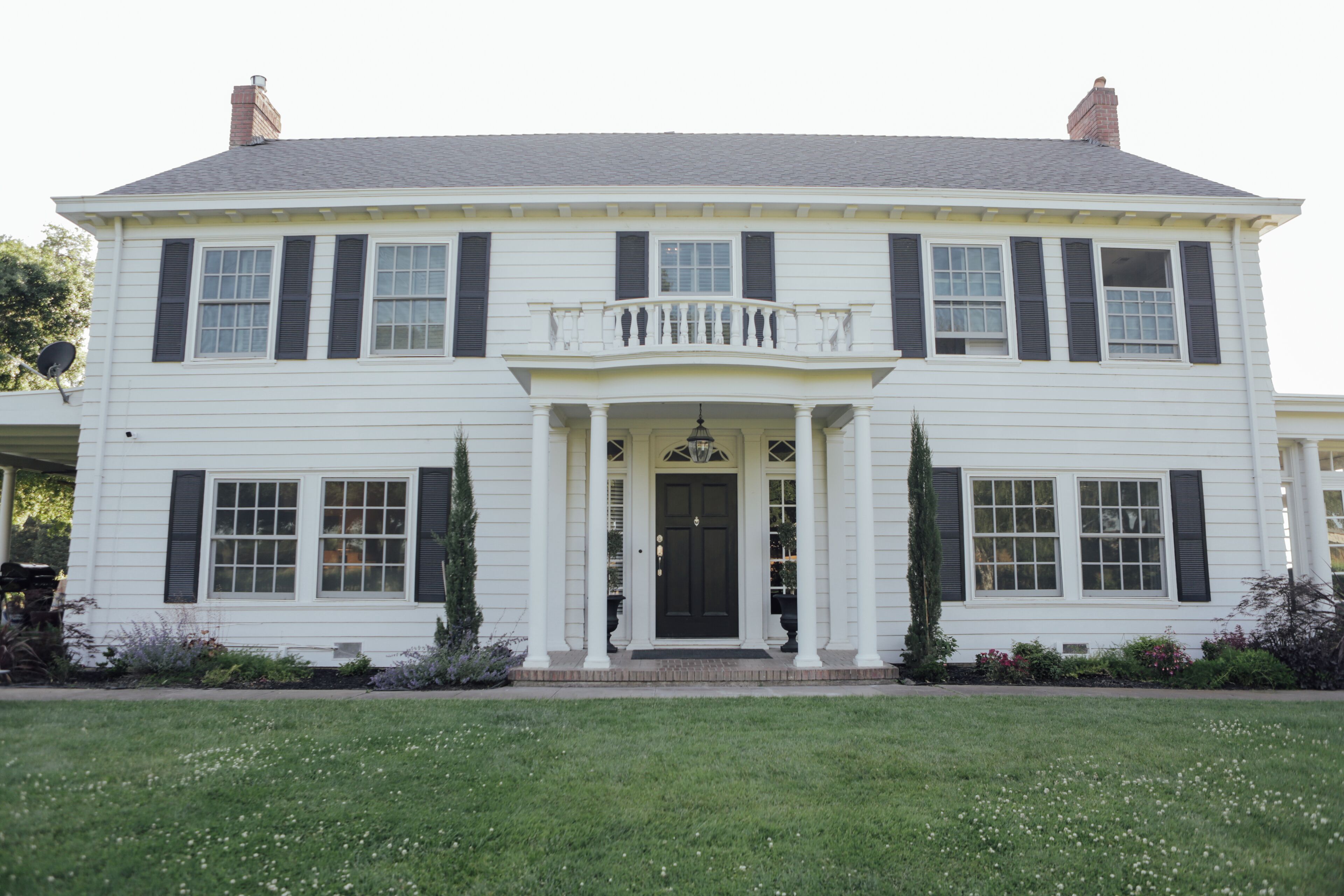 Une maison blanche à deux étages, avec des volets noirs, une entrée centrale surmontée d’un balcon et une pelouse bien entretenue.