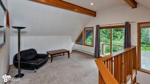 A loft-style room with a sloped ceiling, a black leather sofa, a small wooden table, and a floor lamp. Large windows offer a view of the outdoors. - Spacious rental for 6 in lovely home with gorgeous views, minutes from resort (Girdwood)
