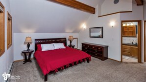 A bedroom with a large bed, wooden headboard, two bedside tables with lamps, a dresser, and a kitchen area visible through an open doorway.