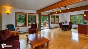 A spacious living area with a wooden dining table, red chairs, and a large window offering a view of a deck and greenery. - Spacious rental for 6 in lovely home with gorgeous views, minutes from resort (Girdwood)
