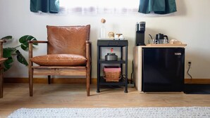 A cozy room with a wooden chair, a small black refrigerator, a coffee maker, and a shelf with kitchen items.