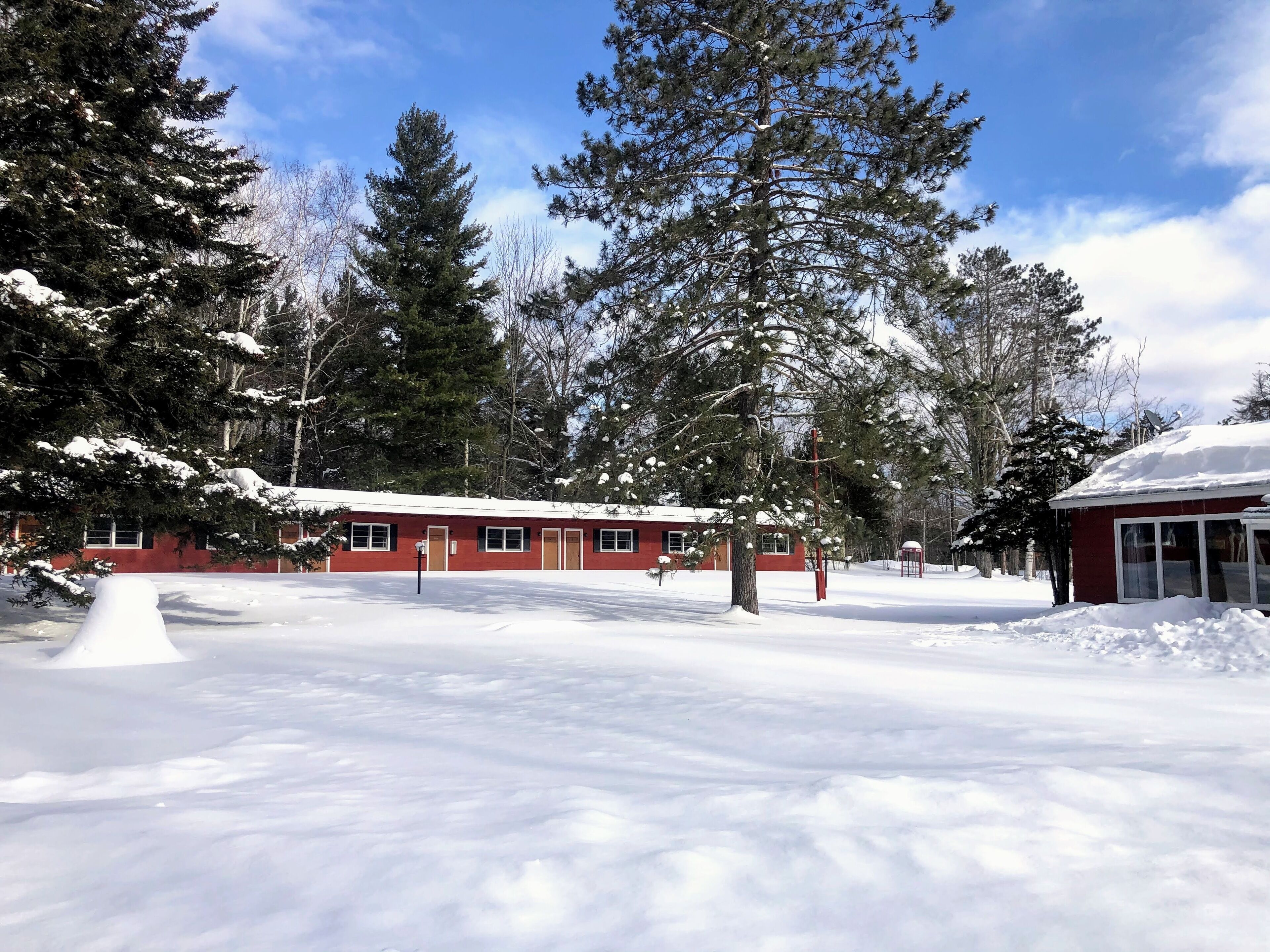 A snow-covered landscape with a red building featuring multiple windows and doors, surrounded by tall evergreen trees.