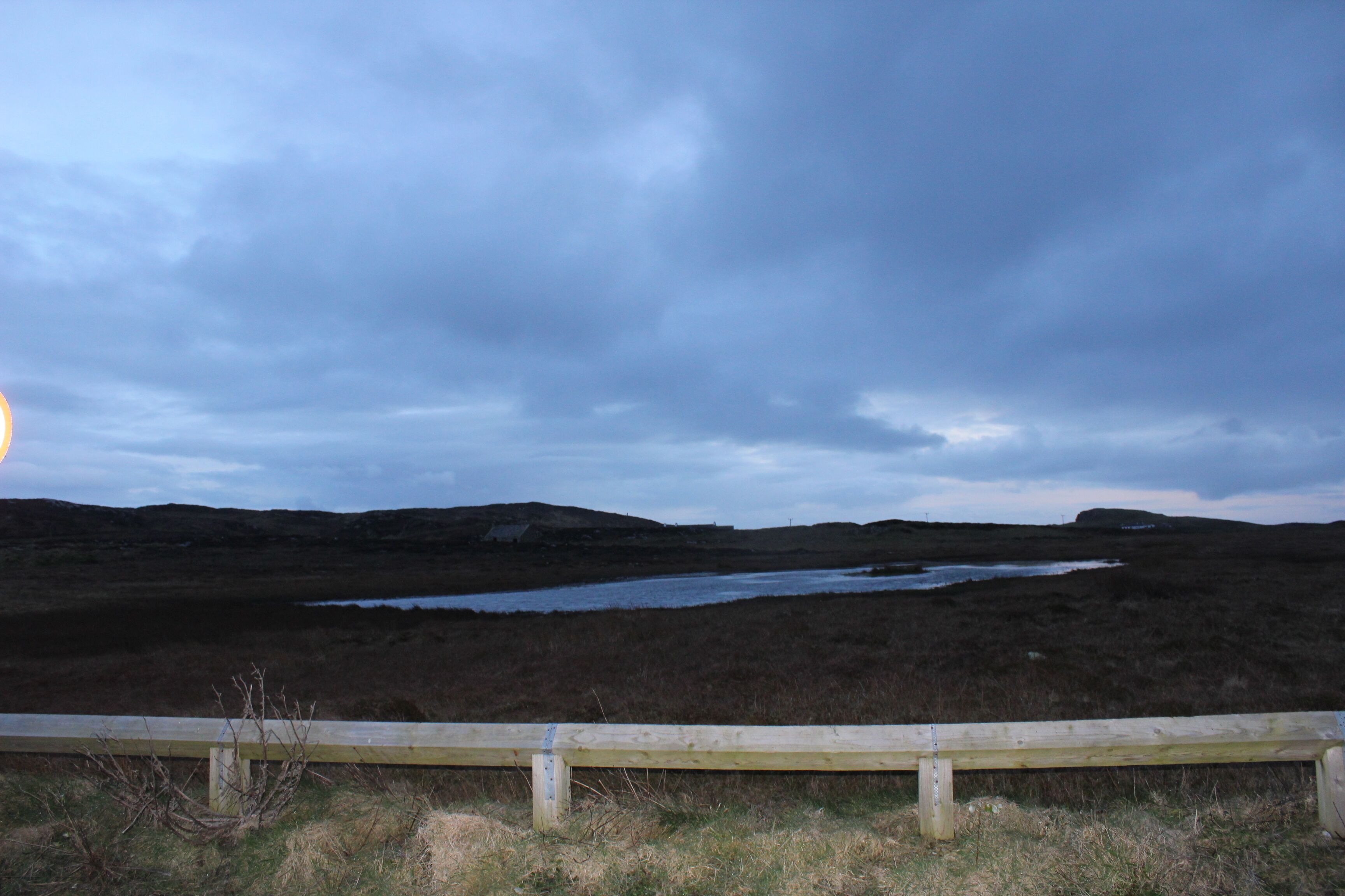 A coastal landscape with a wooden fence, a small body of water, and distant hills.