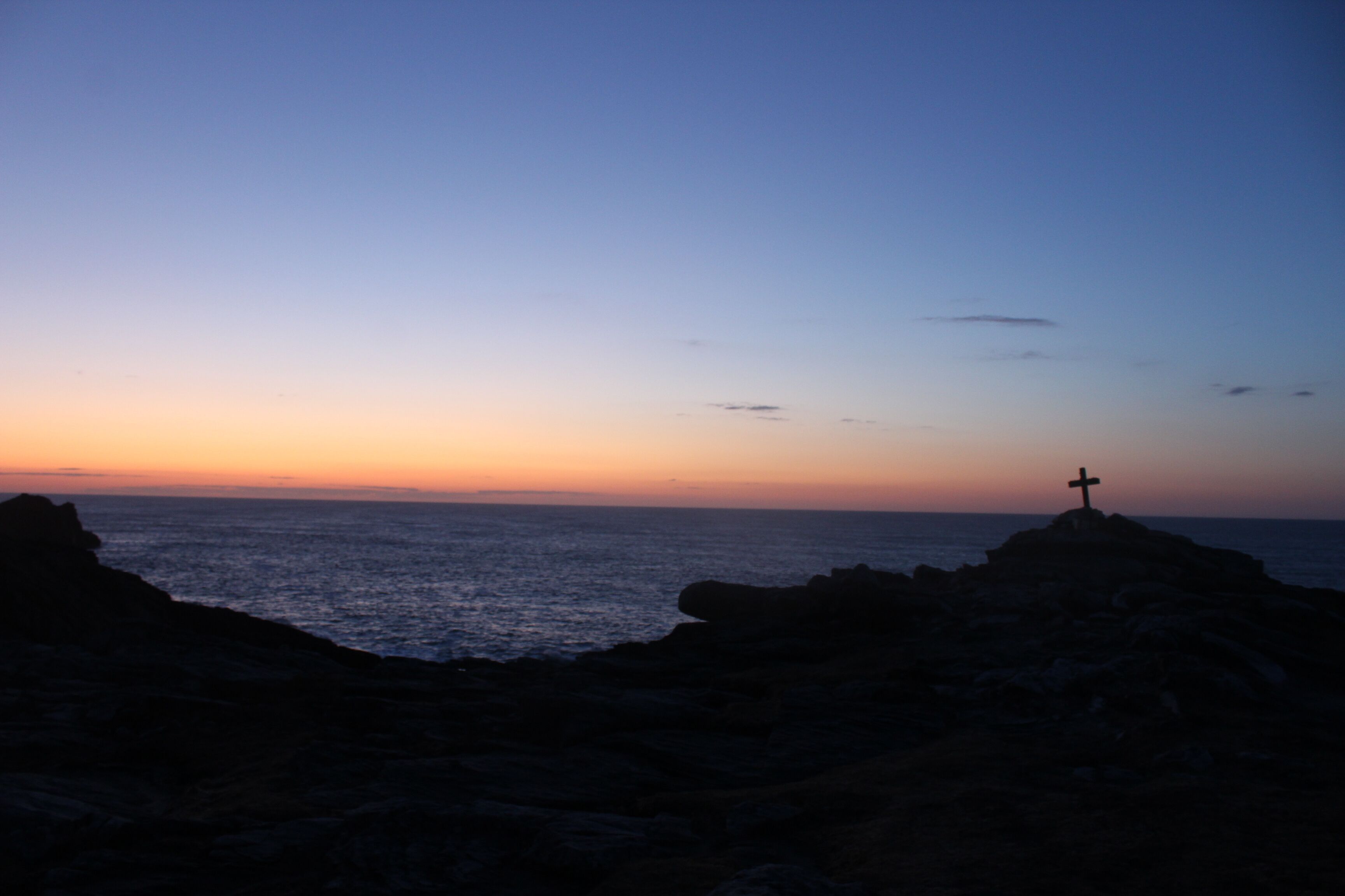 A cross on a rocky outcrop overlooking the sea at sunset.