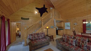 Interno di una baita con una scala in legno, due divani con cuscini decorati, un tavolo da pranzo e una testa di animale appesa al muro.