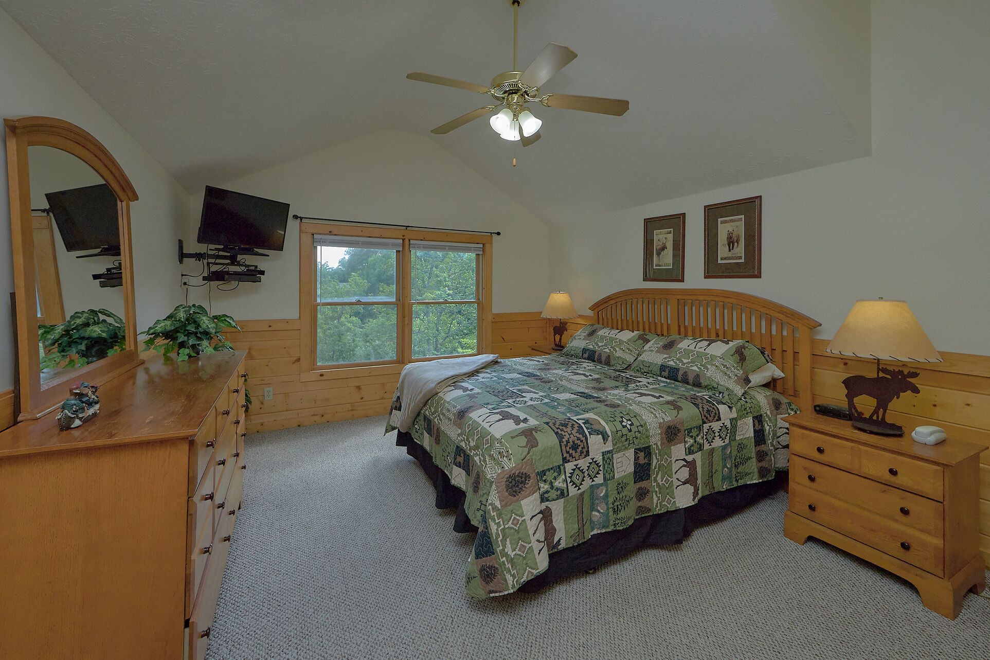 A bedroom with a bed, a ceiling fan, a dresser, a television, and a window with a view of trees.