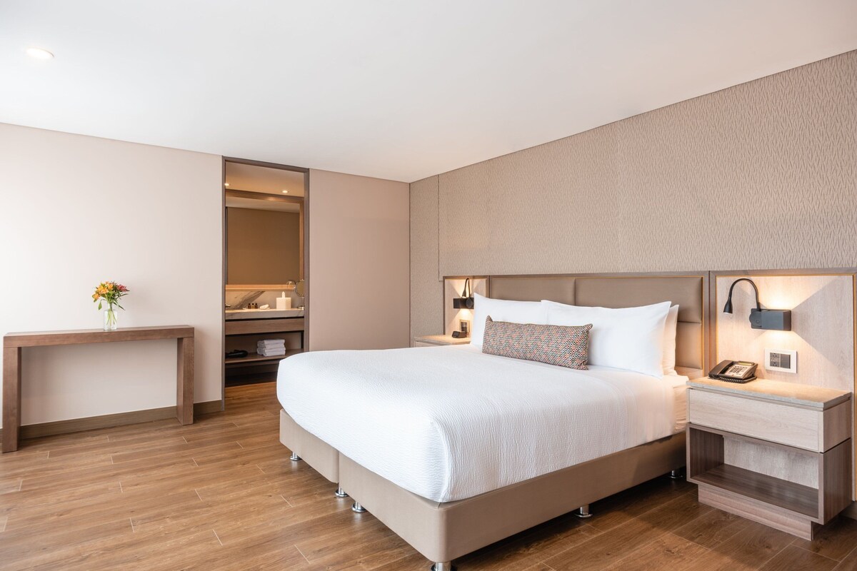 A modern hotel room with a large bed, bedside tables, a nightstand, and a small desk.