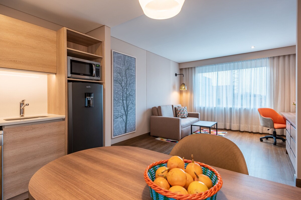 A modern hotel room with a wooden dining table, a microwave, a refrigerator, a sofa, a chair, and a large window with curtains.