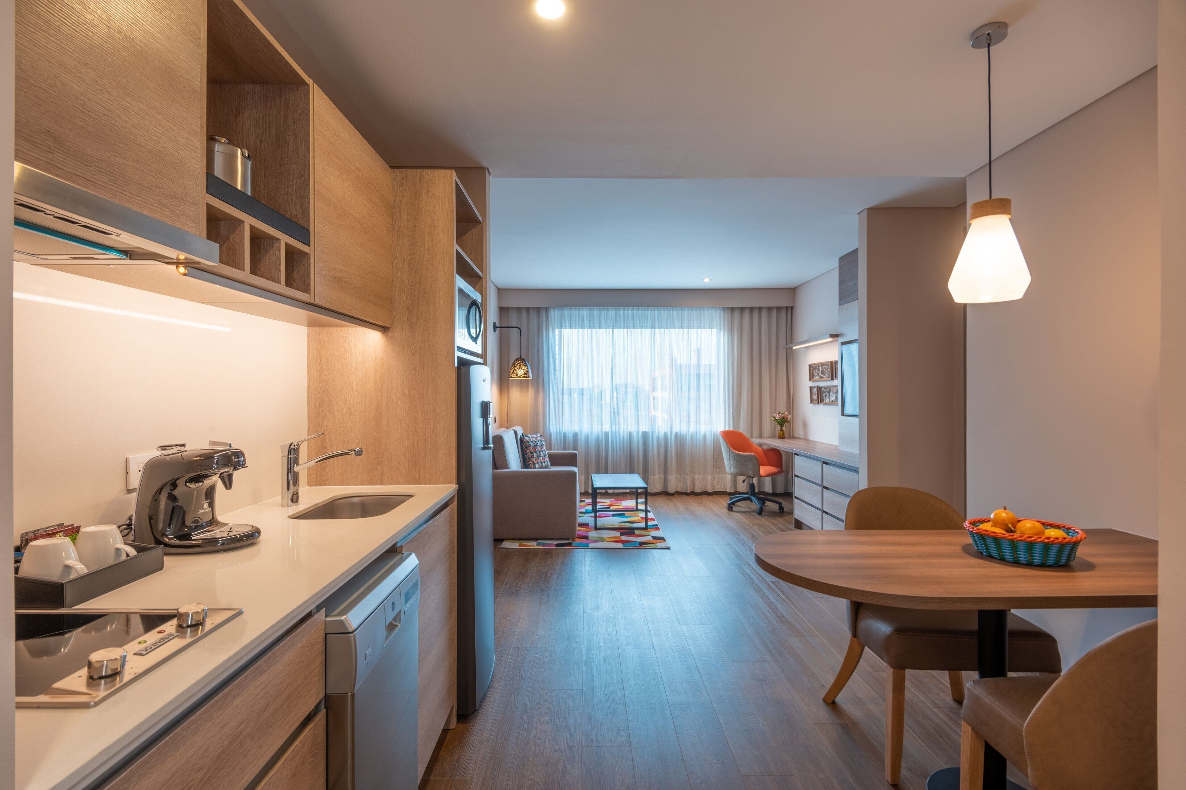 A modern kitchen with a coffee machine, a sink, and a dishwasher. There is a dining area with a table and chairs, and a living room with a sofa and a coffee table.