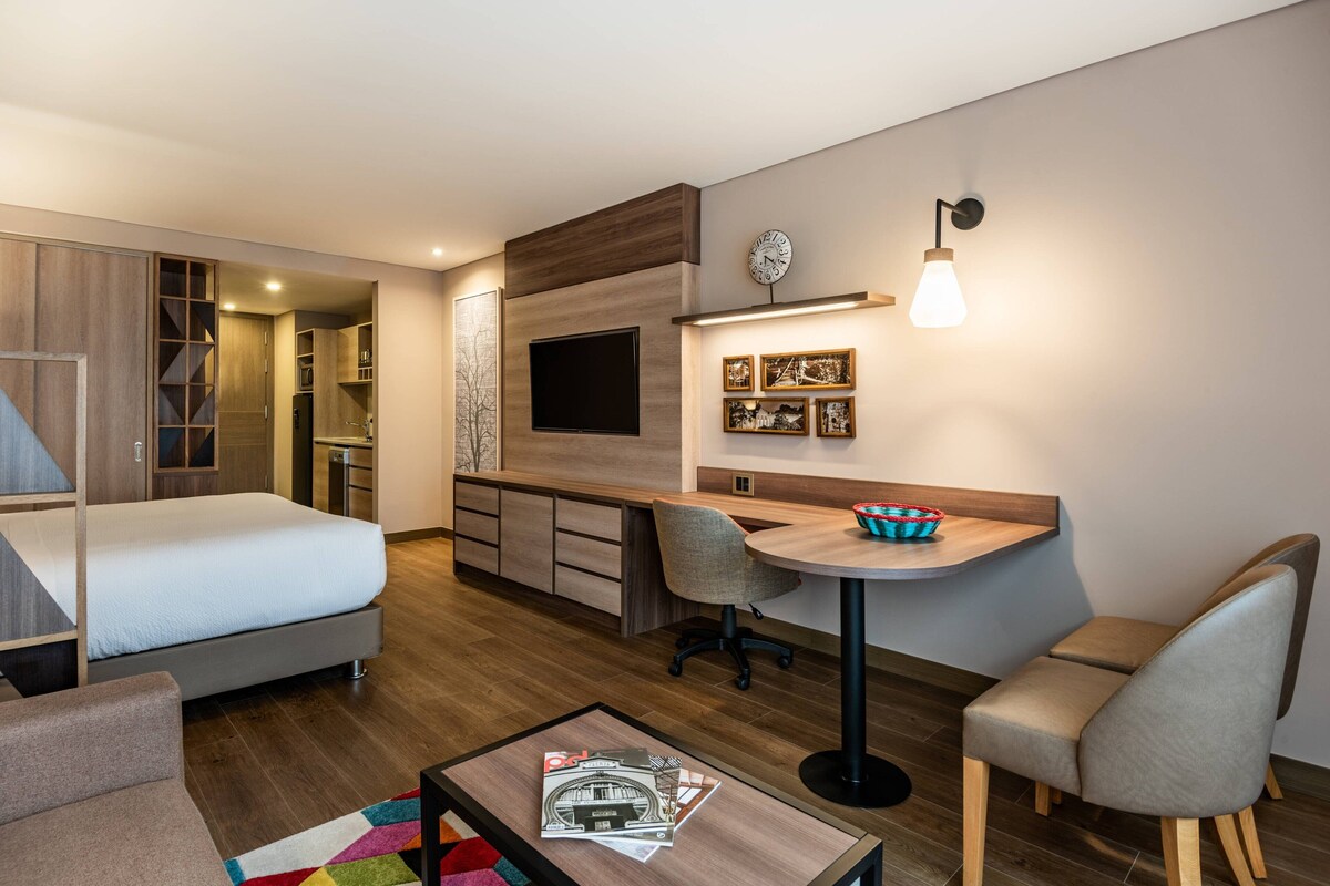 A modern hotel room with a bed, a desk, a chair, a small table, and a TV.
