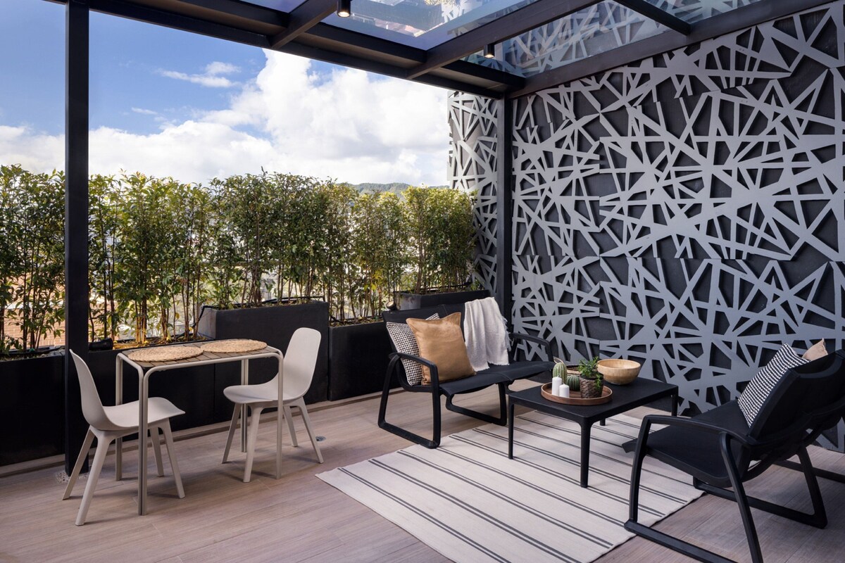 An outdoor patio with a modern design, featuring a geometric wall pattern, a dining table with chairs, and a seating area with cushions.