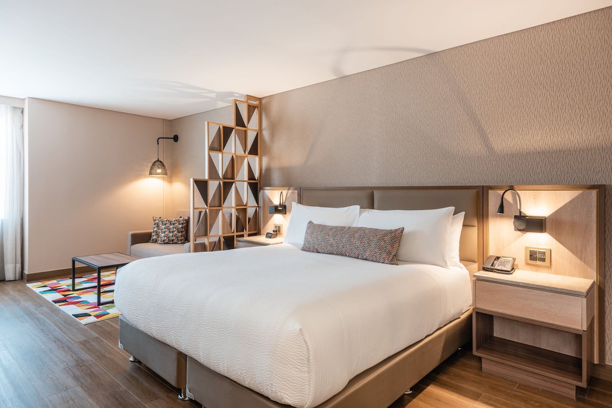 Ett modernt hotellrum med en stor säng, ett träfärgat sänggavel, sängbord med lampor, en mönstrad matta och ett geometriskt mönster på väggen.