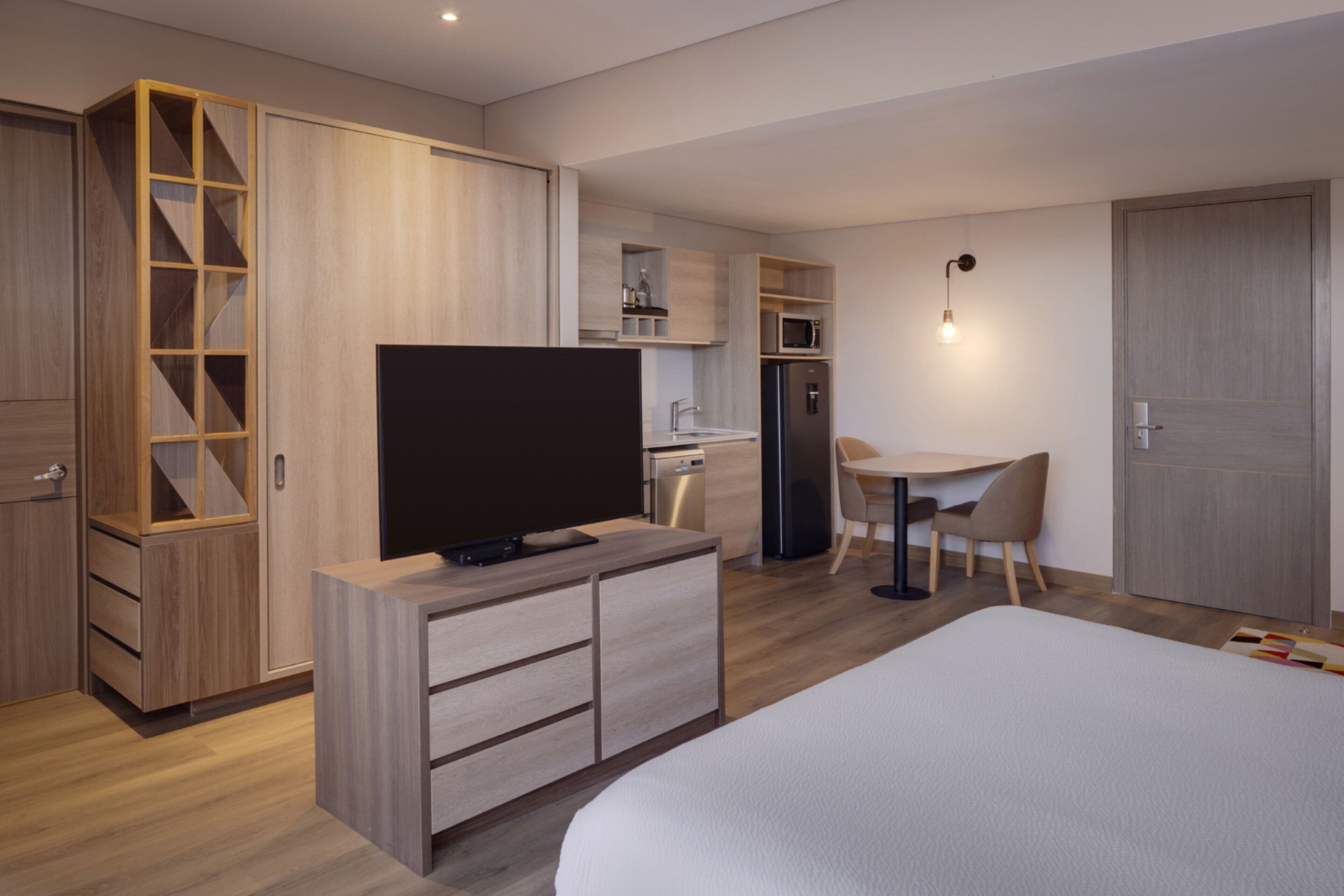 Ein modernes Hotelzimmer mit einem großen Bett, einem Fernseher, einem Schreibtisch mit Stuhl, einer Küchenzeile und einem Kleiderschrank.