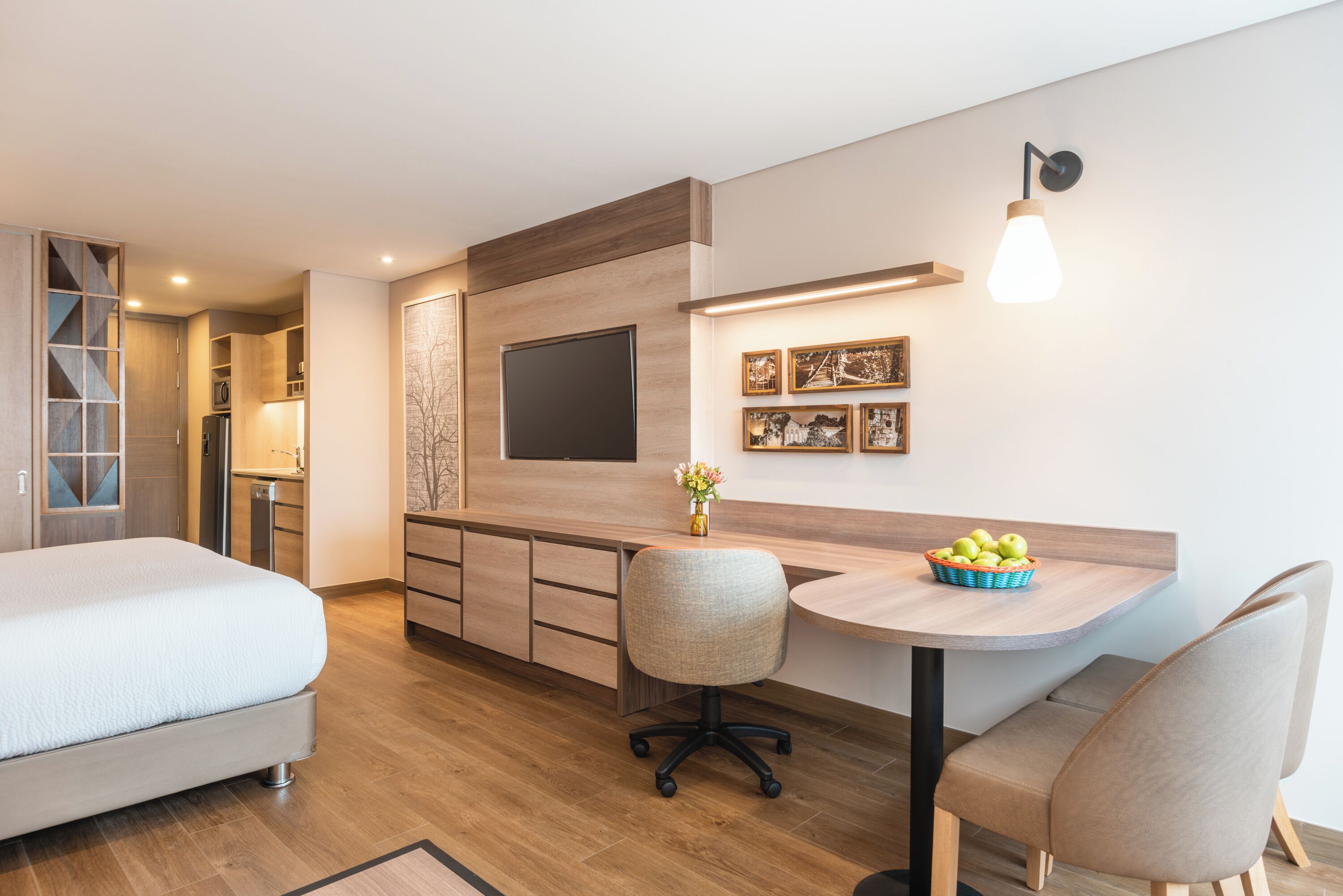 A modern hotel room with a bed, a desk with a chair, a TV, and a wall-mounted light fixture.
