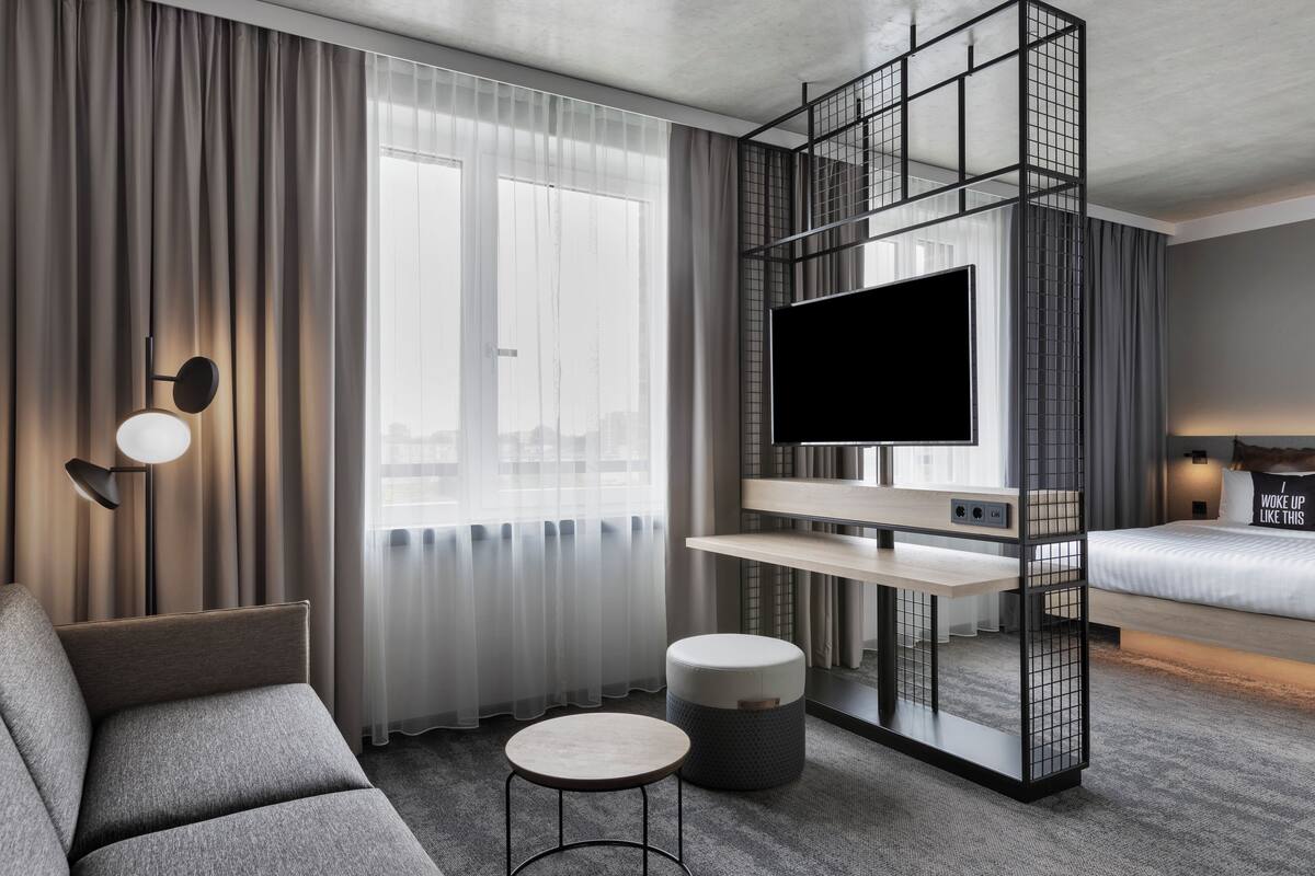 Una habitación de hotel moderna con sofá, una pequeña mesa redonda, televisor sobre un mueble bajo y cama con cabecera.