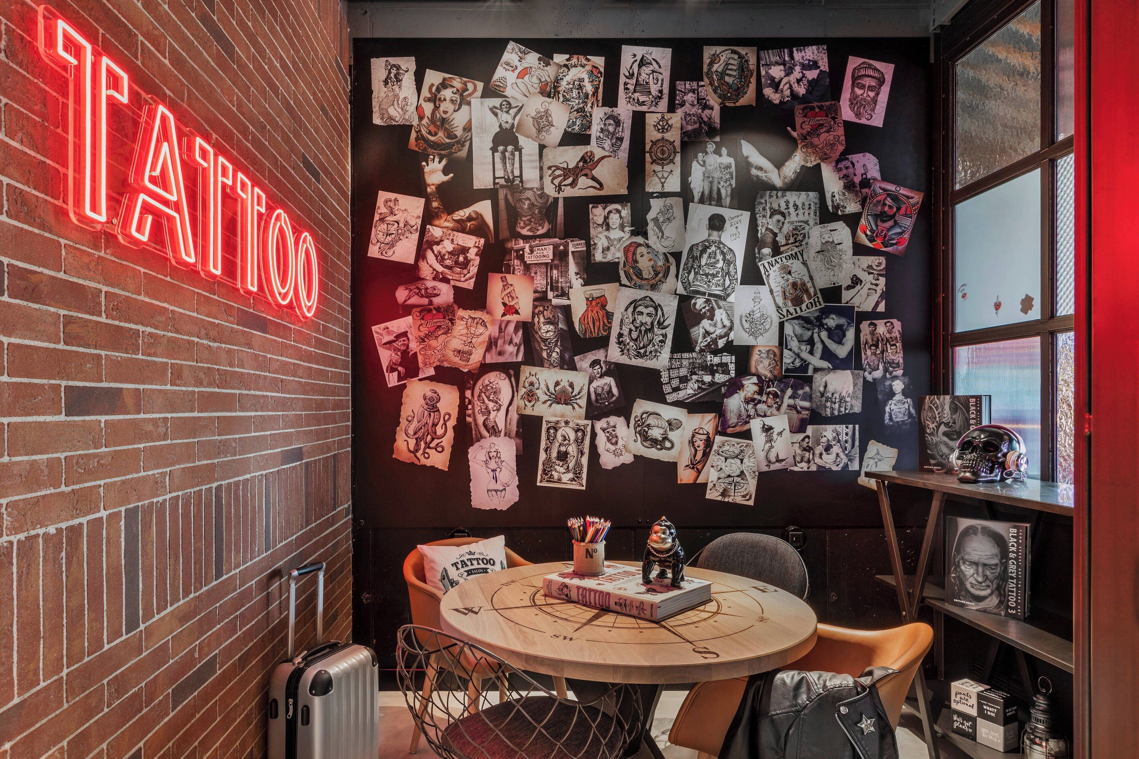 Una habitación con un letrero de neón que dice 'TATTOO', una pared decorada con diseños de tatuajes, una mesa redonda con sillas y una maleta.