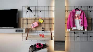 Una habitación moderna con un televisor de pantalla plana, un mueble en la pared para guardar bolsos y una mesita con bebidas, y un espejo que refleja la habitación.