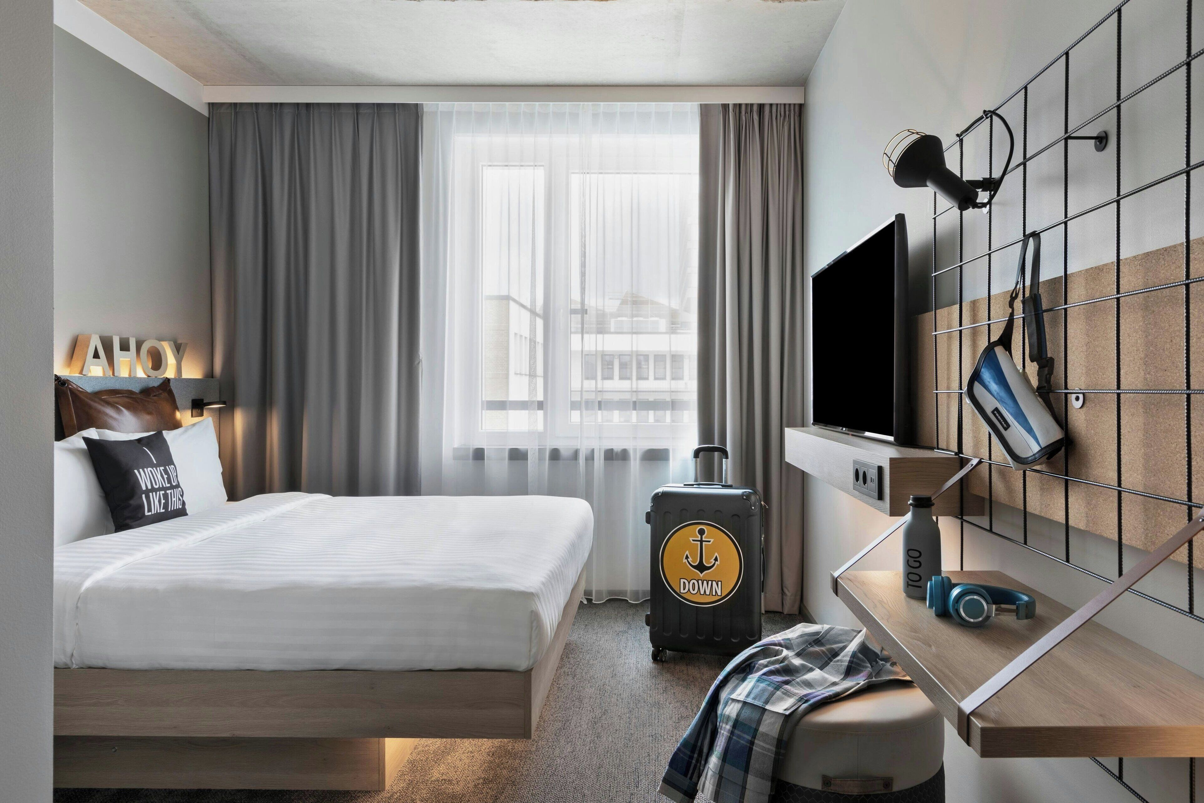 A hotel room with a bed, a desk with a computer, a suitcase, and a TV mounted on the wall.
