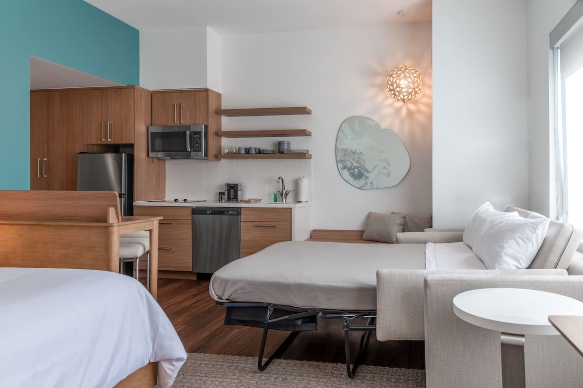Quarto de hotel moderno com cama, sofá, pequena mesa de jantar, área de cozinha com micro-ondas e geladeira, e uma obra de arte única na parede.